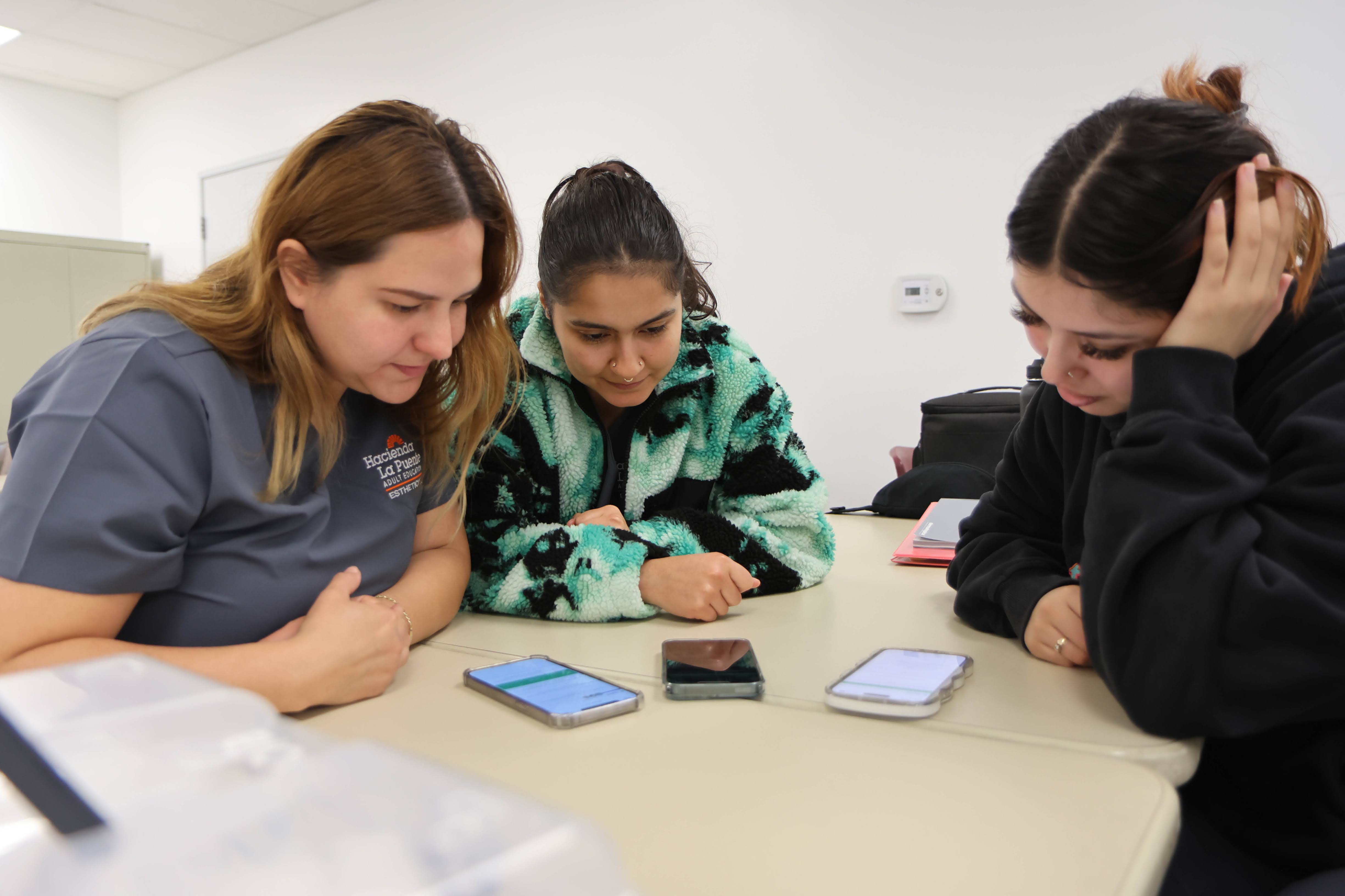 Three women sitting at a table with their heads down, looking at three cellphones.