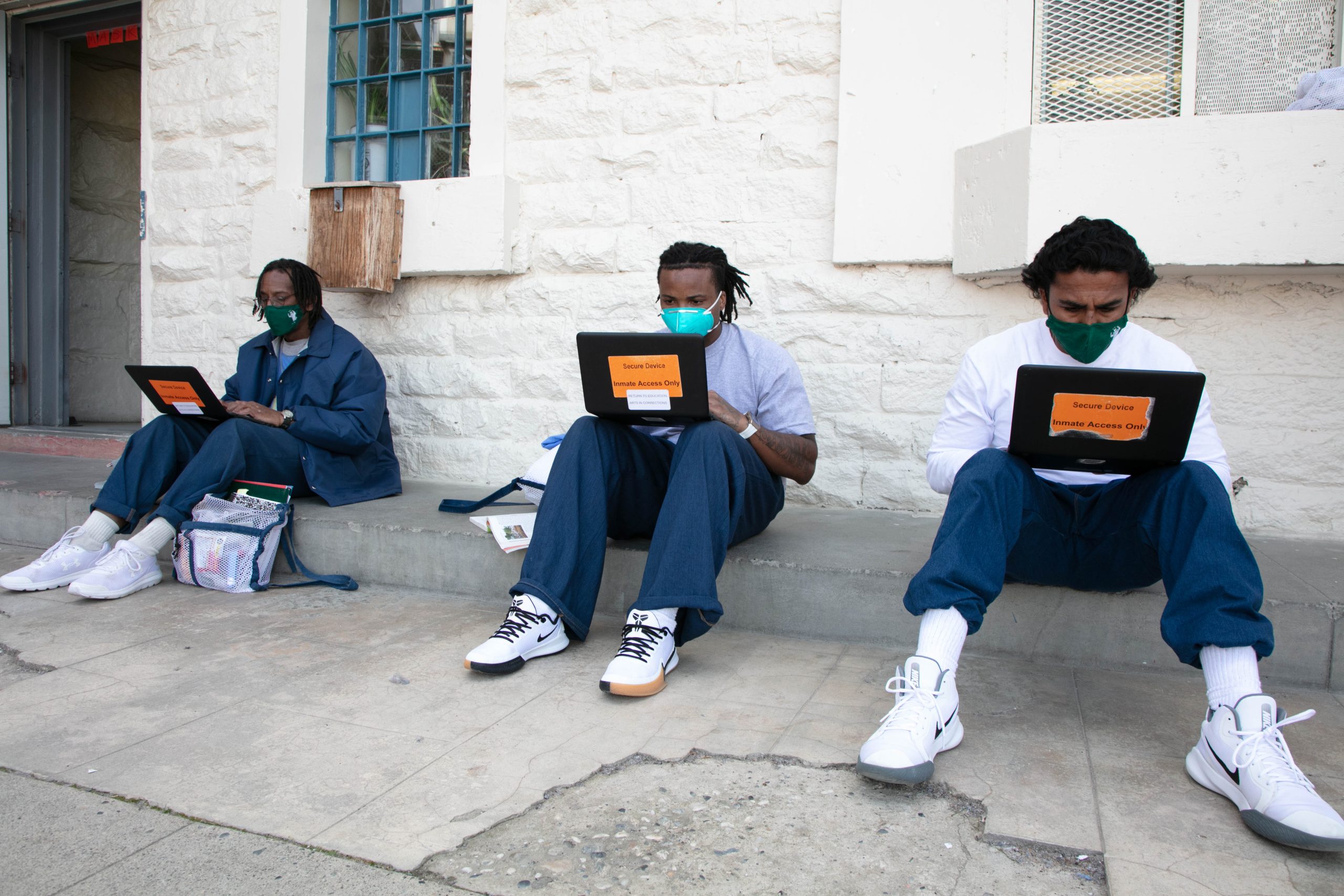 Three incarcerated men sit against a brick wall while using laptops.